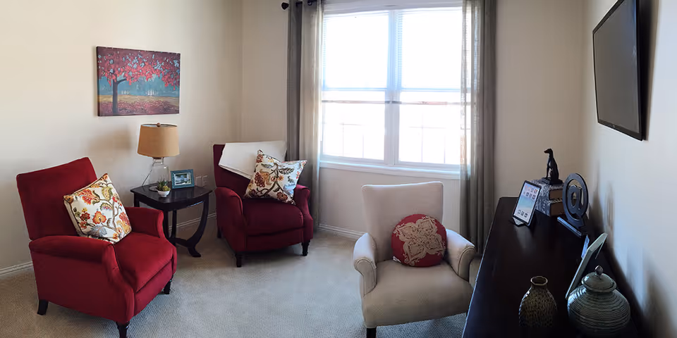 A cozy living room with two red armchairs and one beige armchair, each with decorative pillows. A side table with a lamp and small decor sits between the red chairs. A large window with sheer curtains lets in natural light. A dark wooden console table holds various decorative items and a mounted flat-screen TV is on the wall above it. A colorful painting of a tree hangs on the wall above one of the red chairs.
