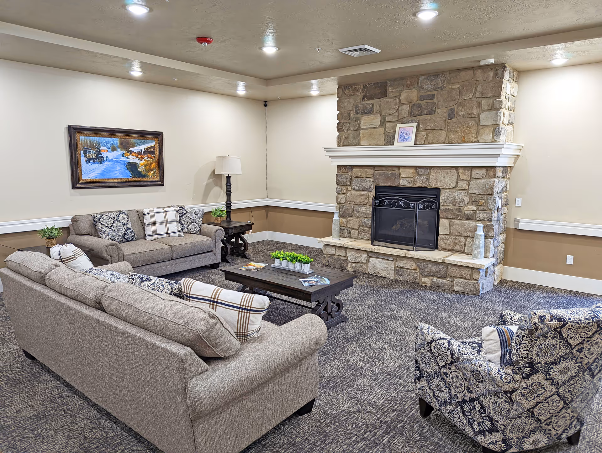 A cozy senior living facility common area with two beige sofas, a patterned armchair, a dark wooden coffee table with small green plants, a stone fireplace with decorative vases, a floor lamp, and a framed painting on the wall.