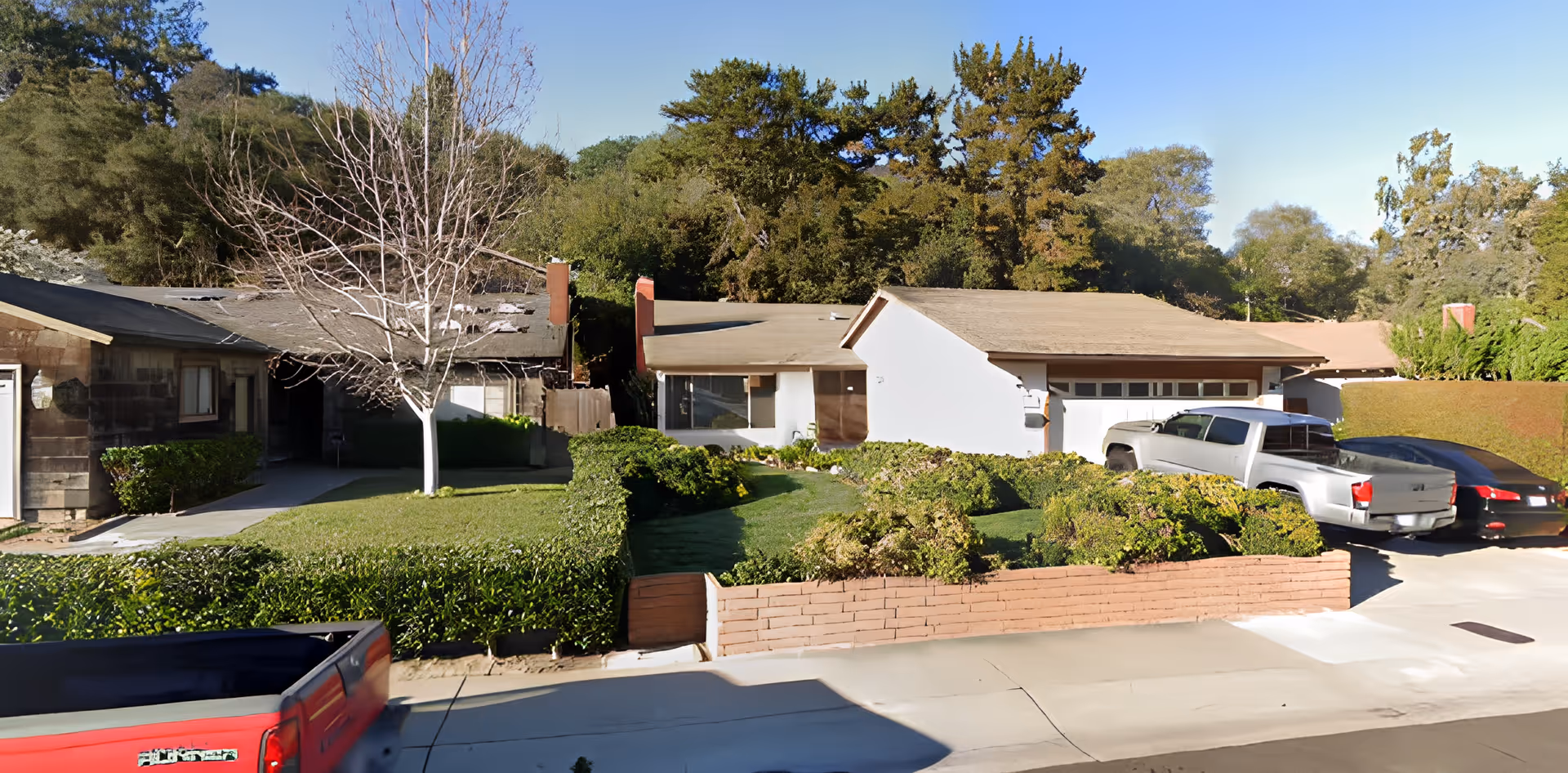 Front exterior view of a residential facility with a driveway, parked vehicles, manicured bushes, a leafless tree, and surrounded by tall trees under a clear blue sky.