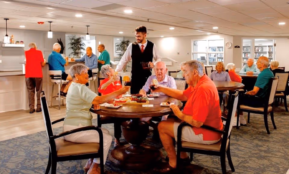 A group of elderly people sitting around tables in a well-lit common dining area, enjoying drinks and snacks. A waiter in a black vest and red tie is serving drinks. Other elderly individuals are seated or standing near a counter in the background, engaging in conversation. The room has a cozy and welcoming atmosphere with carpeted floors and large windows.
