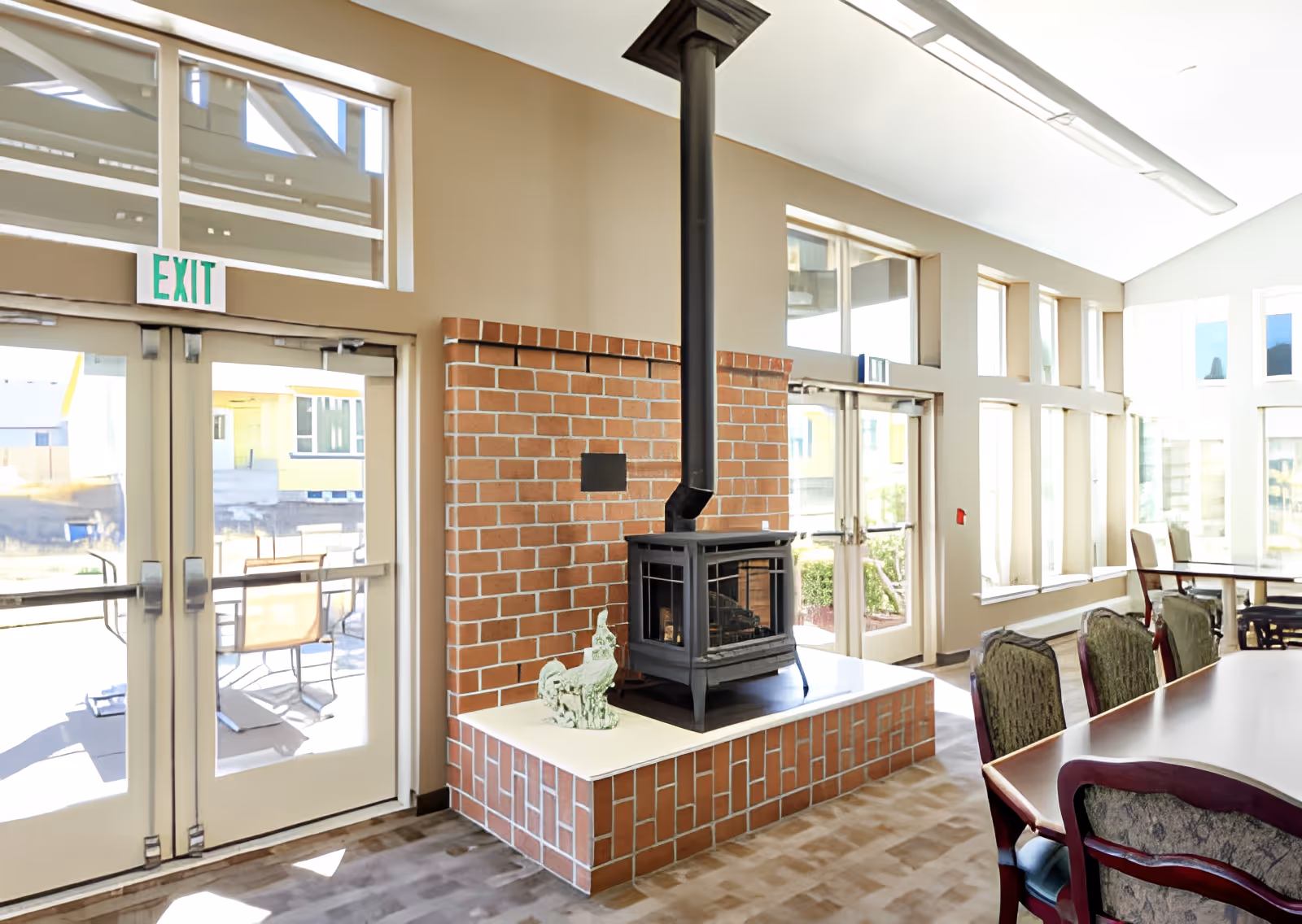 A bright common area with large windows and glass doors letting in natural light. There is a brick fireplace with a black wood stove and a decorative sculpture on the hearth. Several upholstered chairs are arranged around a long table, and outside the glass doors, patio furniture is visible.
