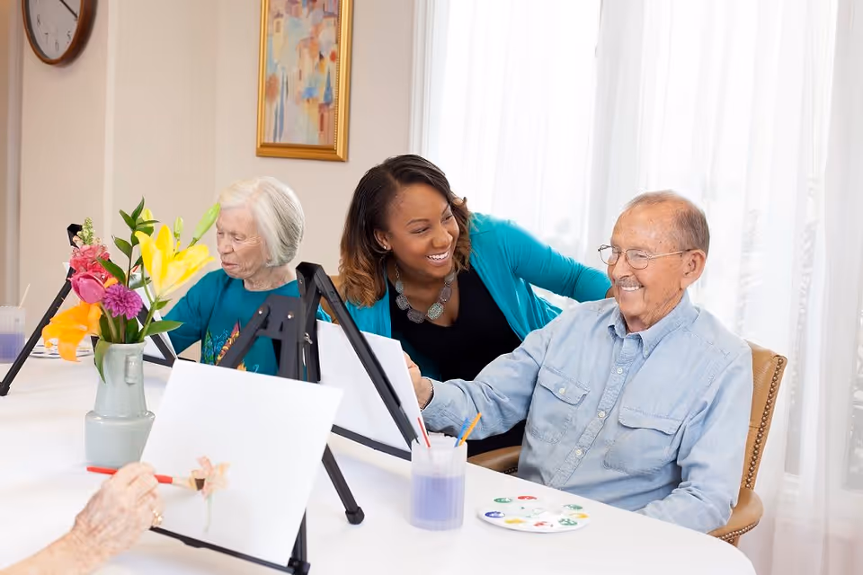 Two elderly individuals seated at a table painting on canvases with a caregiver smiling and interacting with them in a bright room with a clock and framed artwork on the wall.