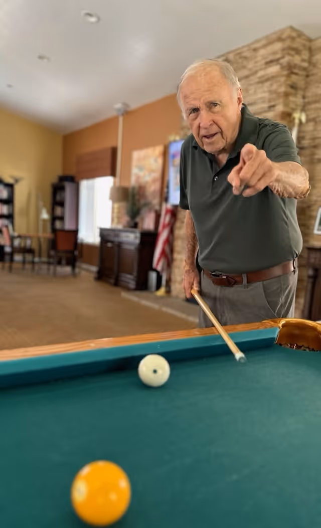An elderly man playing pool indoors, holding a cue stick and pointing towards the camera, with a pool table and balls in the foreground and a cozy room with furniture and decorations in the background.