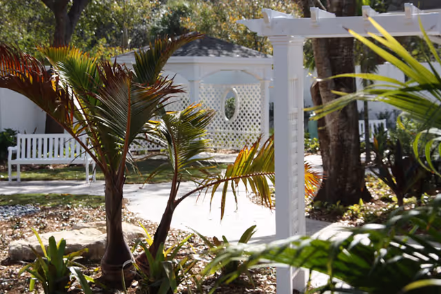 A peaceful outdoor garden area with tropical plants, a white wooden bench, a white pergola, and a white gazebo with lattice walls and a circular window, surrounded by trees and greenery.