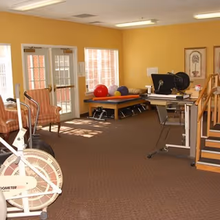 A therapy or exercise room with yellow walls and brown carpet. The room contains exercise equipment including a stationary bike, therapy balls on a table, a treadmill, chairs, and a set of stairs for physical therapy. There are windows and a glass door letting in natural light.