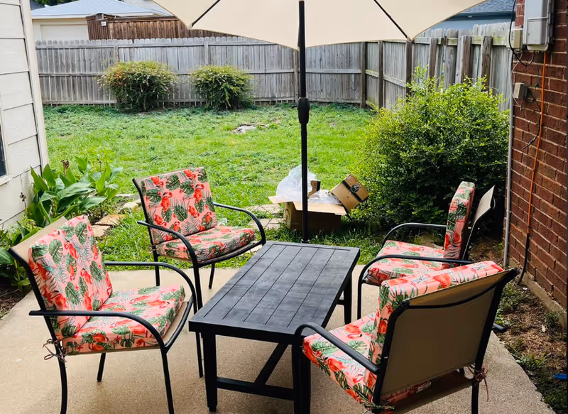 Outdoor patio area with a black rectangular table and five chairs with floral patterned cushions arranged around it. A large umbrella is positioned in the center of the table. The patio is adjacent to a grassy backyard enclosed by a wooden fence, with bushes and plants along the edges.