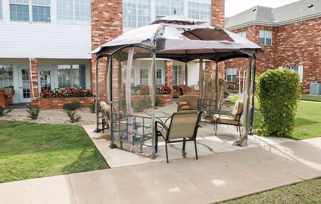 Outdoor patio area with a gazebo featuring a canopy and mesh sides, surrounded by several cushioned chairs. The patio is part of a brick building with windows and flower beds visible in the background.