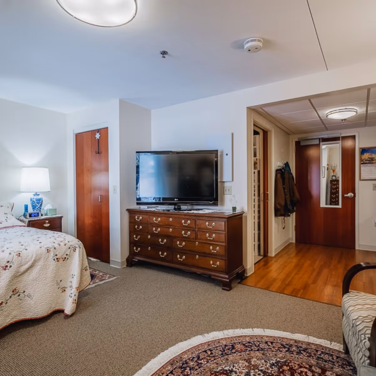 A cozy bedroom in a senior living facility featuring a bed with a floral quilt, a wooden dresser with a flat-screen TV on top, a nightstand with a lamp, and a wooden door with a mirror. The room has carpeted flooring with a round patterned rug and a small sitting area with a cushioned chair.