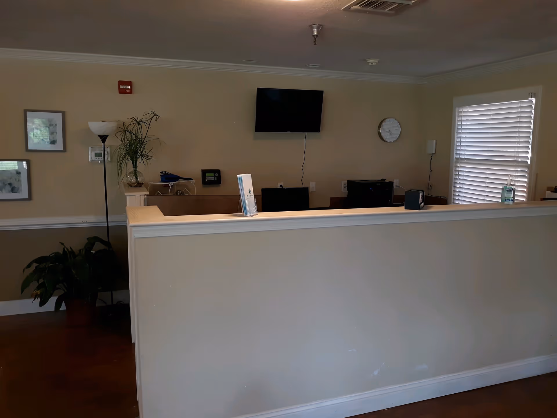 Reception area with a beige half-wall counter, a flat-screen TV mounted on the wall, a clock, a floor lamp, two framed pictures, a potted plant, and a window with white blinds. There is a brochure holder on the counter and a bottle of hand sanitizer near the window.