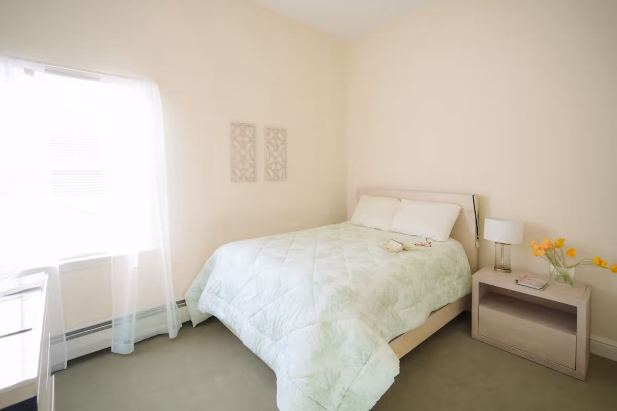 A bright bedroom with a bed covered in a light green patterned comforter and two white pillows. There is a nightstand next to the bed with a lamp, a book, and a vase with yellow flowers. Two decorative wall hangings are above the bed, and a window with sheer white curtains lets in natural light.
