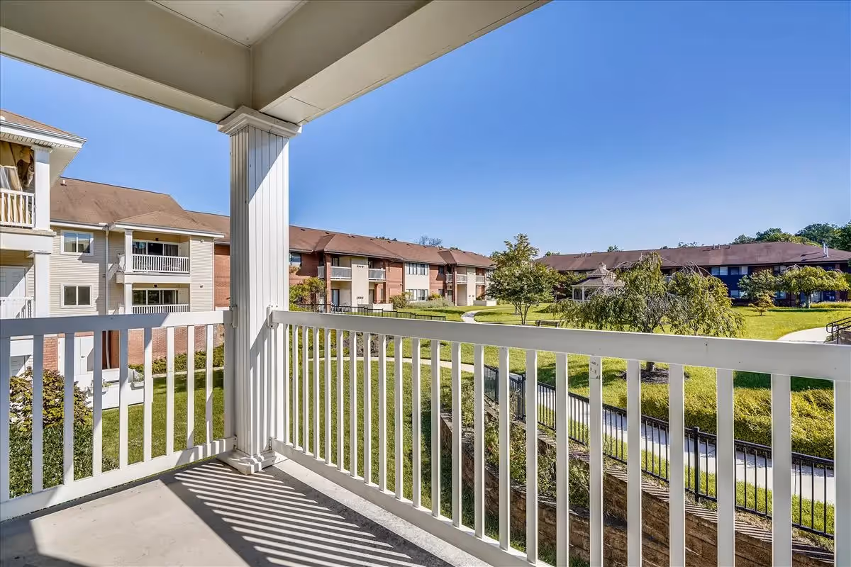 View from a covered balcony with white railing overlooking a green courtyard with trees and walking paths, surrounded by multi-story residential buildings under a clear blue sky.