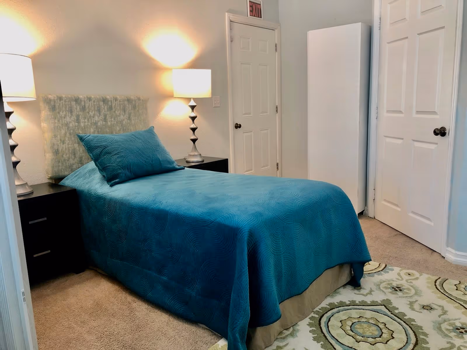 A bedroom with a single bed covered in a teal bedspread and matching pillow. There are two bedside tables, each with a lamp. The room has beige carpet, a patterned area rug, and white walls with two closed white doors and a white wardrobe.