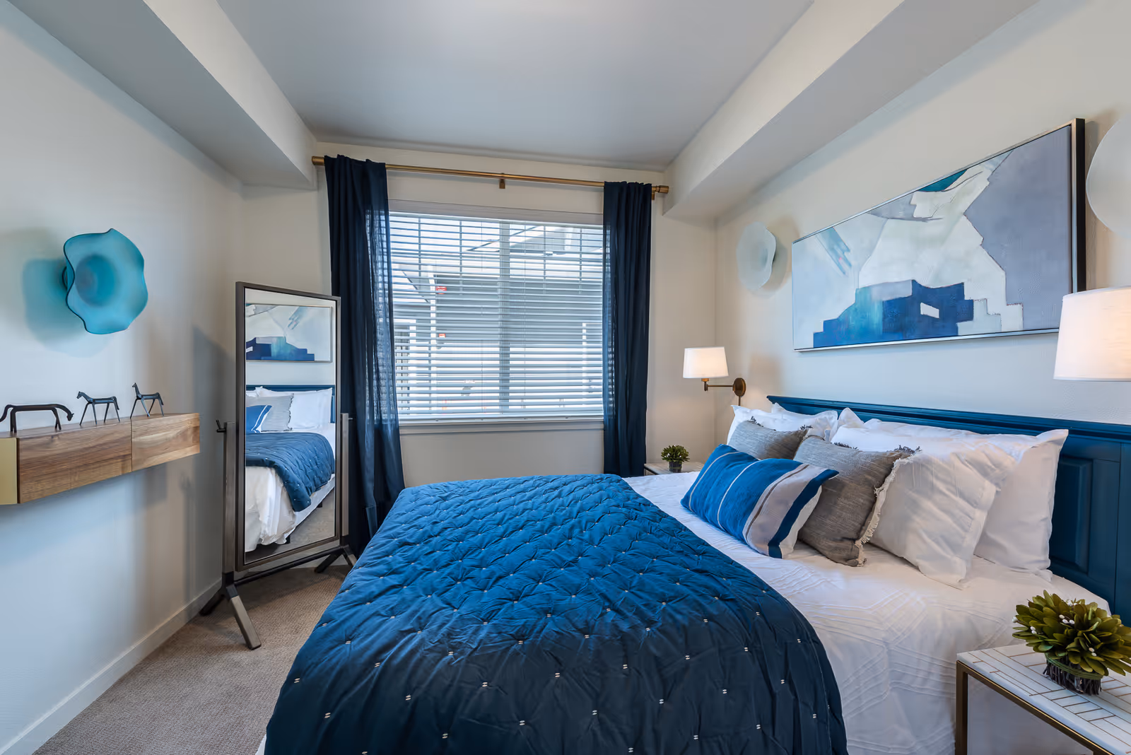 A neatly made bed with a blue quilt and multiple pillows in a bedroom. The room features a large window with blinds and dark blue curtains, a standing mirror reflecting part of the bed, modern wall-mounted lamps, abstract wall art above the bed, and small decorative plants on bedside tables.