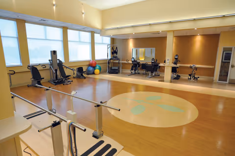 A spacious fitness room with exercise equipment including stationary bikes, weight machines, and colorful exercise balls. The room has large windows letting in natural light, wooden flooring with a circular design in the center, and parallel bars for walking exercises.
