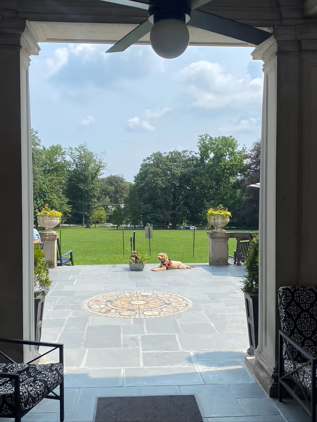 Covered patio opening onto a tiled terrace and green lawn, with a dog lying near a planter under a ceiling fan.