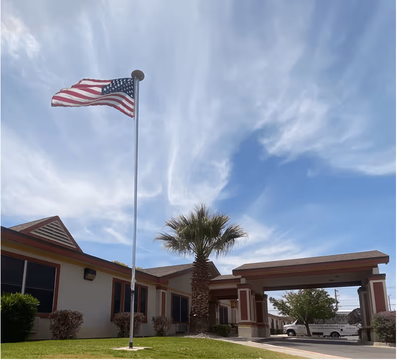 Front entrance of a senior living facility with an American flag on a tall pole, a palm tree, and a blue sky with wispy clouds.