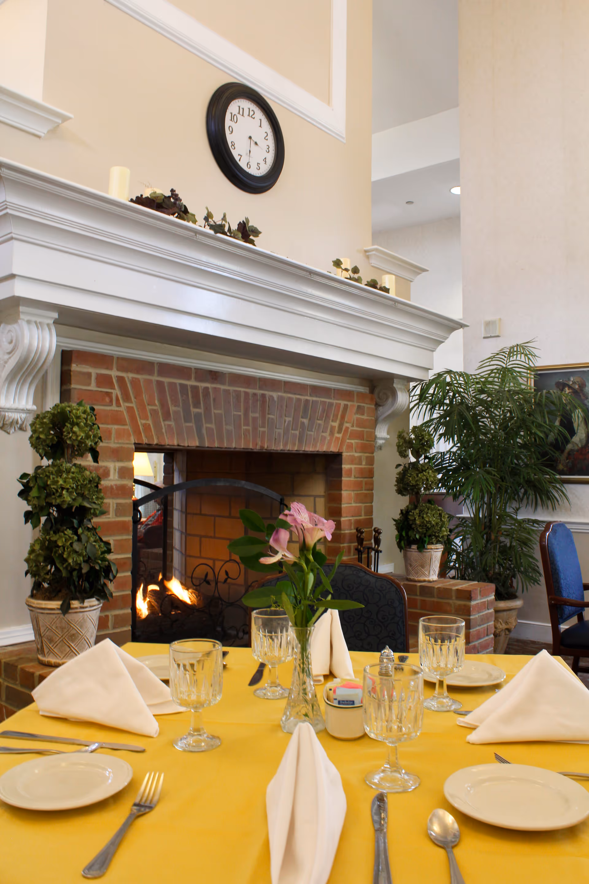 A dining table set with plates, glasses, silverware, and folded white napkins on a yellow tablecloth in front of a brick fireplace with a fire burning. The fireplace has a white mantel decorated with greenery and candles, and a round clock hangs above it. There are potted plants and chairs around the table in a cozy indoor setting.