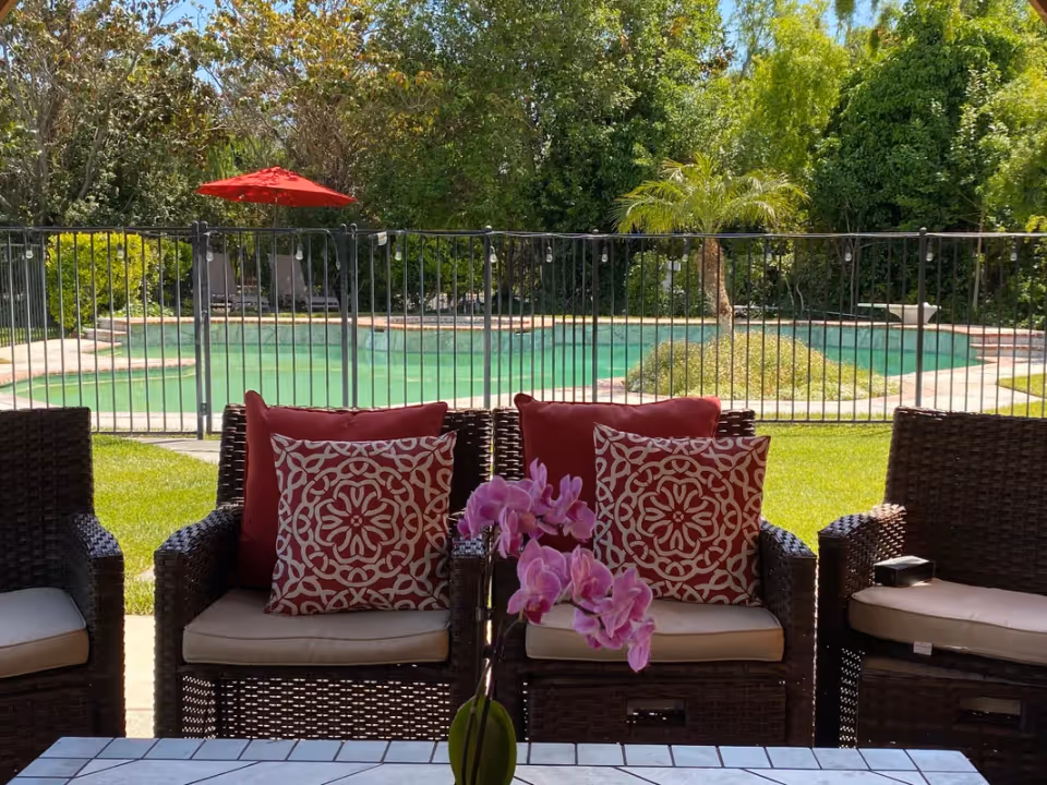 Outdoor seating area with wicker chairs featuring red and white patterned cushions, a tiled table with a pink orchid centerpiece, a fenced swimming pool, green grass, trees, and a red umbrella in the background.