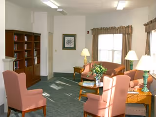 A bright common living room with upholstered chairs and sofas arranged around coffee tables, lamps, a bookshelf, and windows with valances.