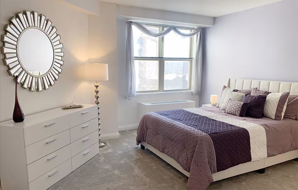 A bright and neatly arranged bedroom featuring a large bed with purple and white bedding and multiple pillows. To the left, there is a white dresser with a decorative vase and a round mirror above it. A tall floor lamp stands between the dresser and the window, which is adorned with sheer curtains allowing natural light to fill the room.