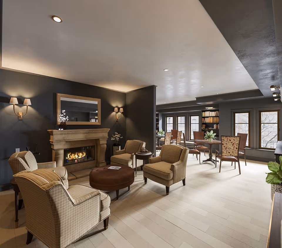 A cozy senior living common area with four upholstered armchairs arranged around a round brown ottoman in front of a stone fireplace with a fire burning. The walls are dark with two wall sconces on either side of a large mirror above the fireplace. In the background, there are tables and chairs near large windows and bookshelves filled with books.