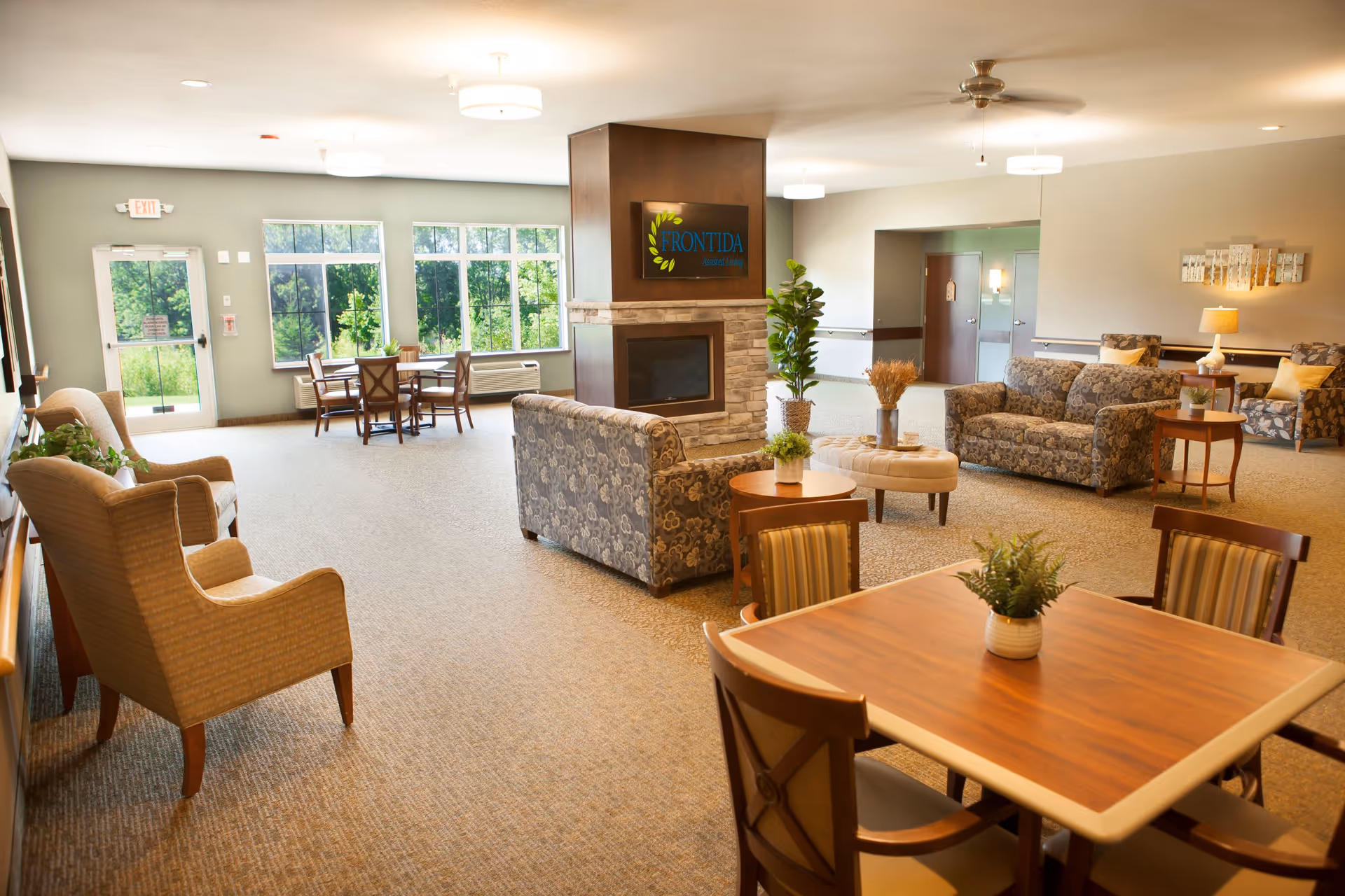 A spacious and well-lit common living area in a senior living facility featuring multiple seating arrangements including armchairs, sofas, and wooden tables with chairs. The room has large windows letting in natural light, a central fireplace with a sign above it that reads 'Frontida,' and decorative plants and lamps enhancing the cozy atmosphere.
