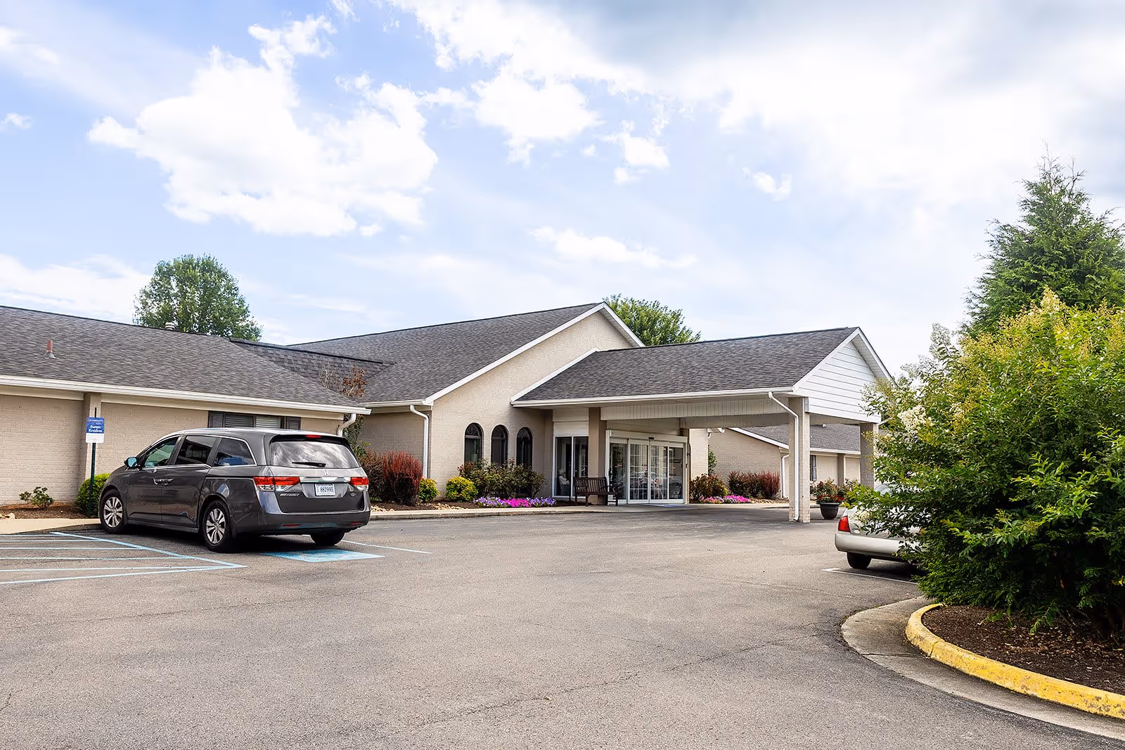 Exterior view of Commonwealth Senior Living at Radford showing the front entrance with a covered drop-off area, a parking lot with a few cars, landscaped bushes, and a partly cloudy sky.