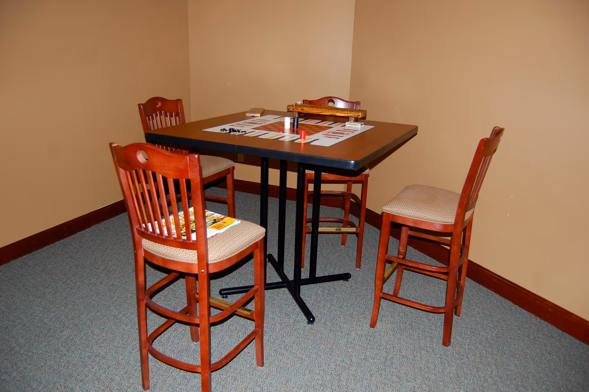 A small square table with a board game set up on it, surrounded by four wooden chairs with cushioned seats, in a room with beige walls and carpeted floor.