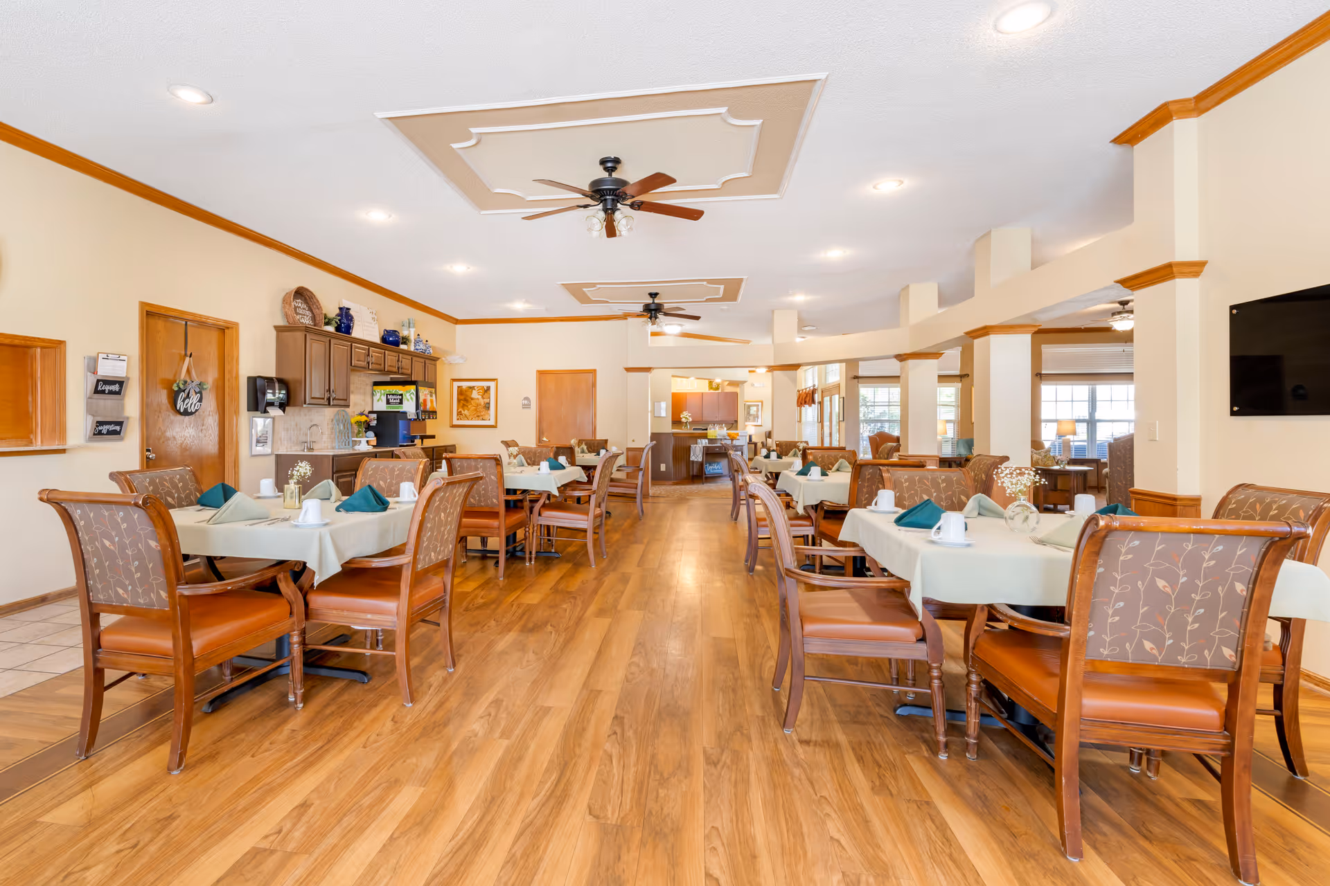 A spacious dining room in a senior living facility with multiple tables covered with light-colored tablecloths, each set with cups, napkins, and small flower vases. The room features wooden chairs with patterned upholstery, ceiling fans, recessed lighting, and wood flooring. There is a beverage station and cabinetry along one wall, and large windows letting in natural light.