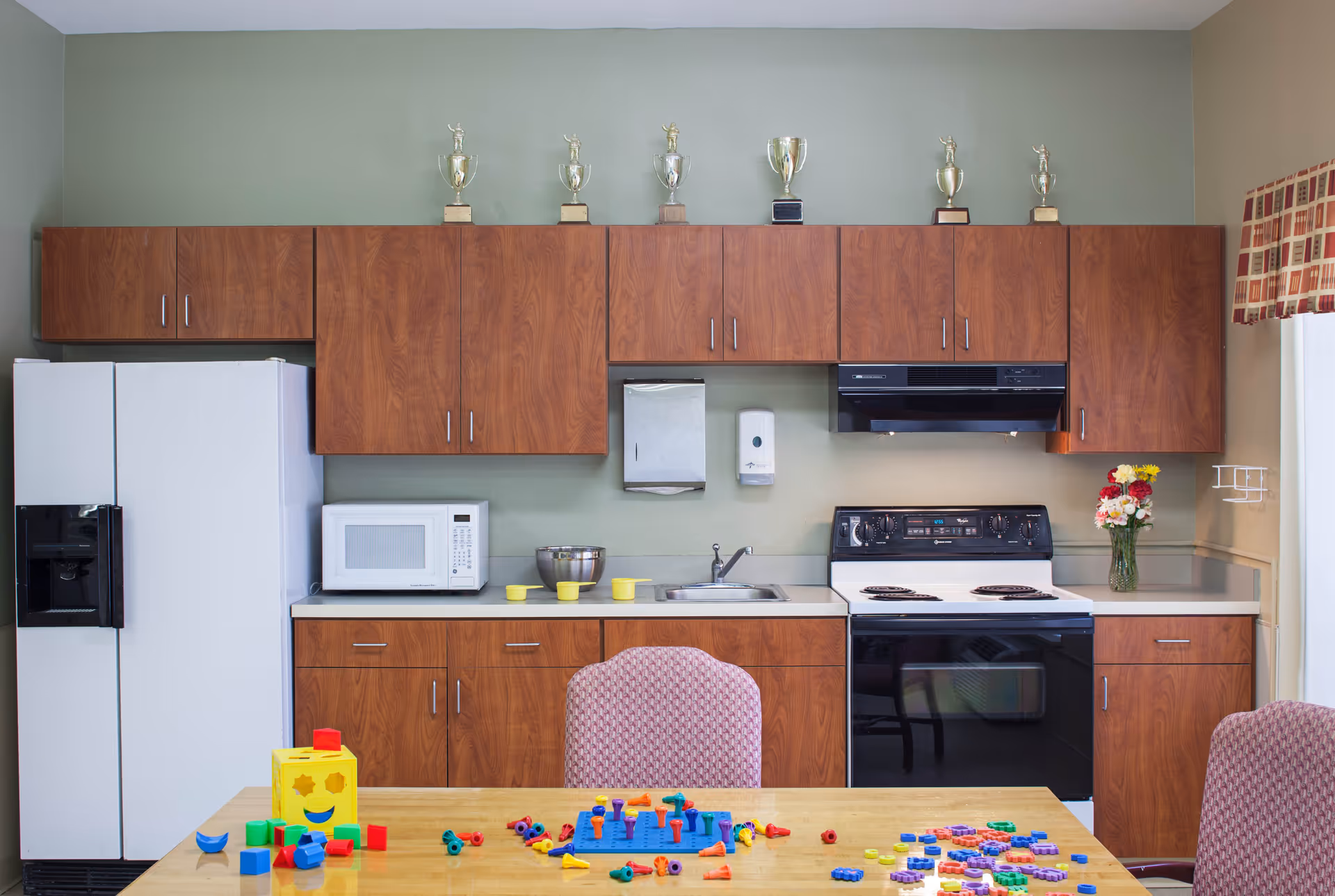 A kitchen area with wooden cabinets, a white refrigerator, microwave, sink, and stove. On the countertop, there is a vase with flowers and some yellow cups. Above the cabinets, there are several trophies displayed. In the foreground, a table with colorful children's toys and two patterned chairs is visible.