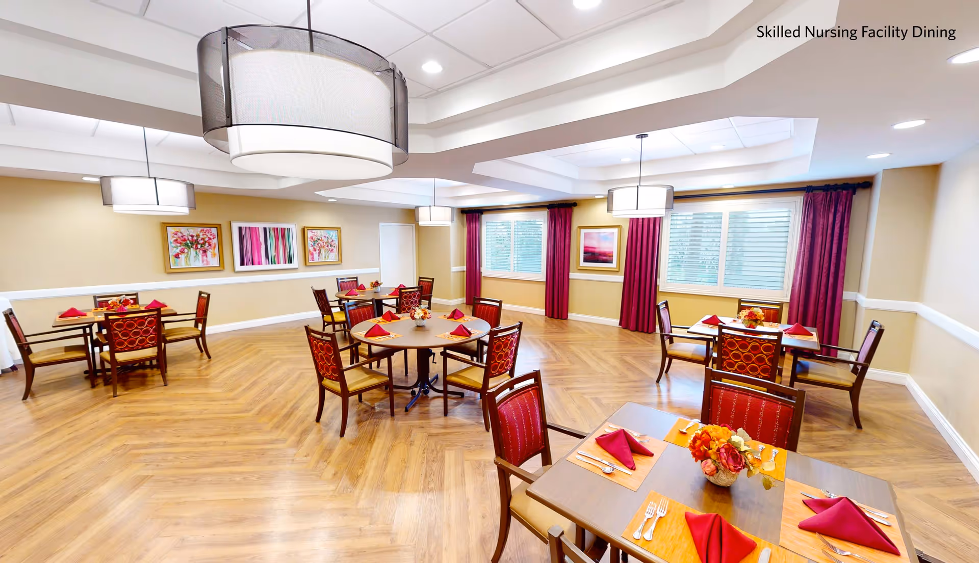 Bright dining room with round and square tables set with red napkins, chairs, pendant lights, artwork, and wood-patterned flooring.