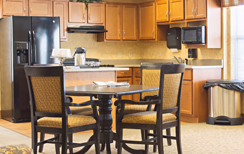 A communal dining area with a round table and four chairs in front of a kitchenette with wooden cabinets, a refrigerator, microwave, and sink.