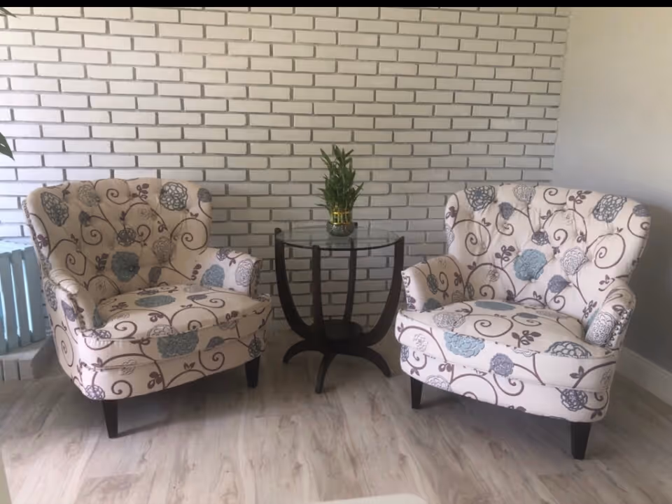 Two upholstered armchairs with floral patterns in shades of blue and gray are positioned on either side of a small round glass-top table with a dark wooden base. A small potted plant is placed on the table. The background features a white brick wall and light-colored wooden flooring.