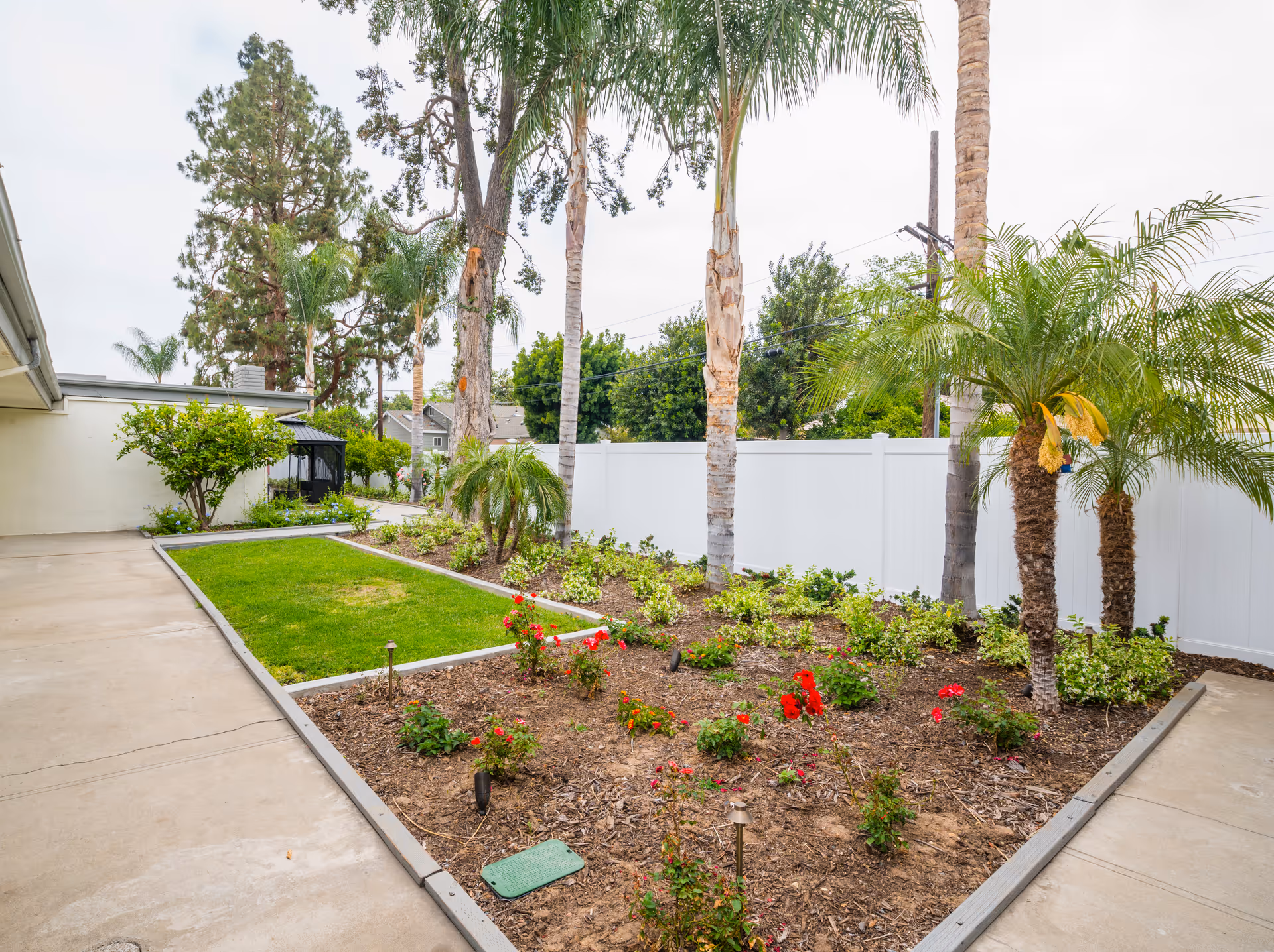 Outdoor garden area at Gordon Lane Care Center featuring a landscaped flower bed with red flowers, small palm trees, and other greenery bordered by a concrete walkway and a white fence in the background.