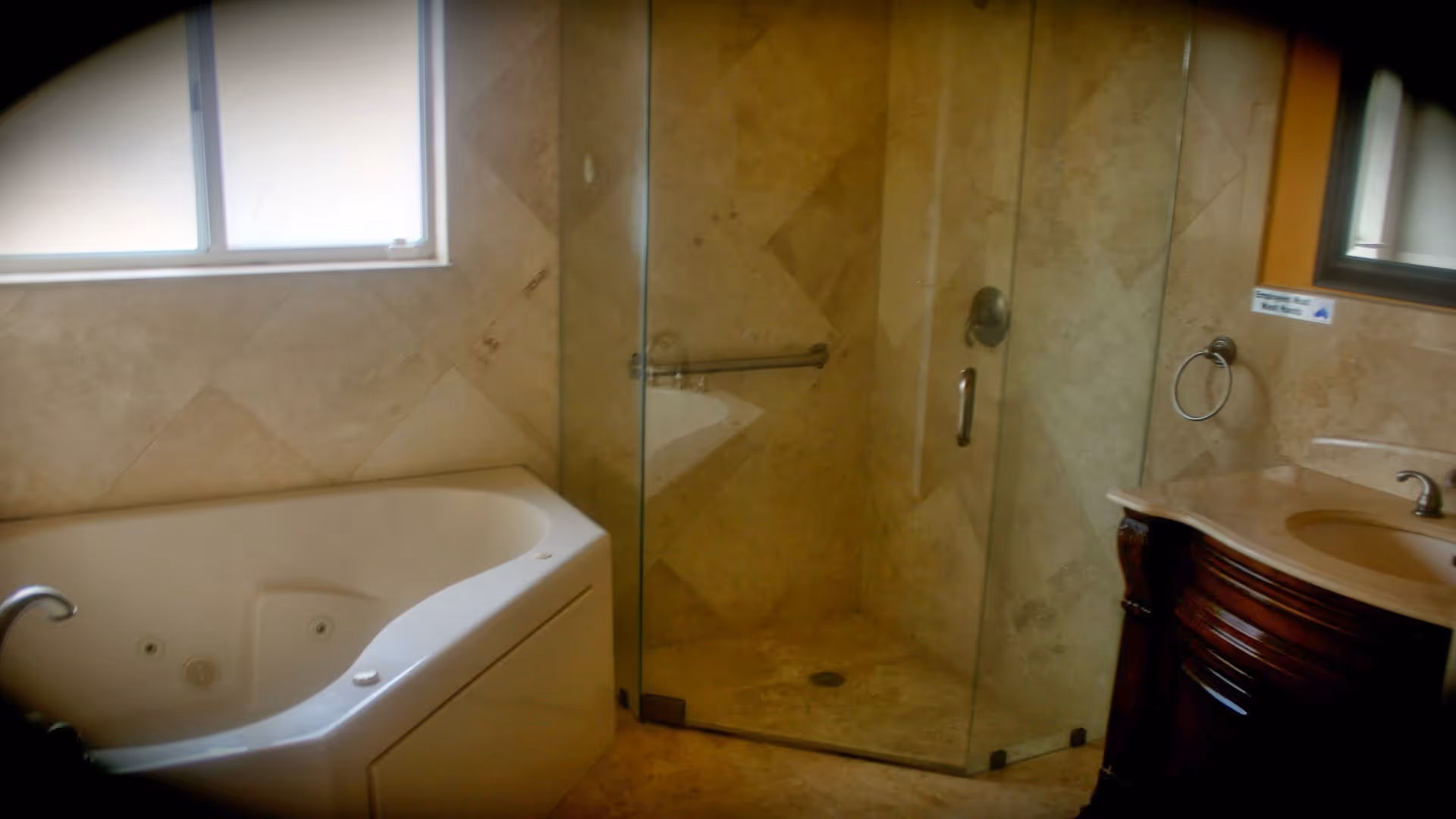 Bathroom with a large white bathtub, a glass-enclosed shower with a metal grab bar, and a wooden vanity with a sink and faucet. The walls and floor are tiled in beige tones, and there is a frosted window above the bathtub.