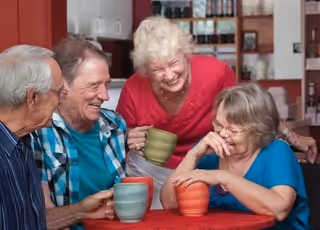 Four elderly people sitting around a table, smiling and holding colorful mugs, enjoying each other's company in a cozy indoor setting with shelves and kitchen items in the background.