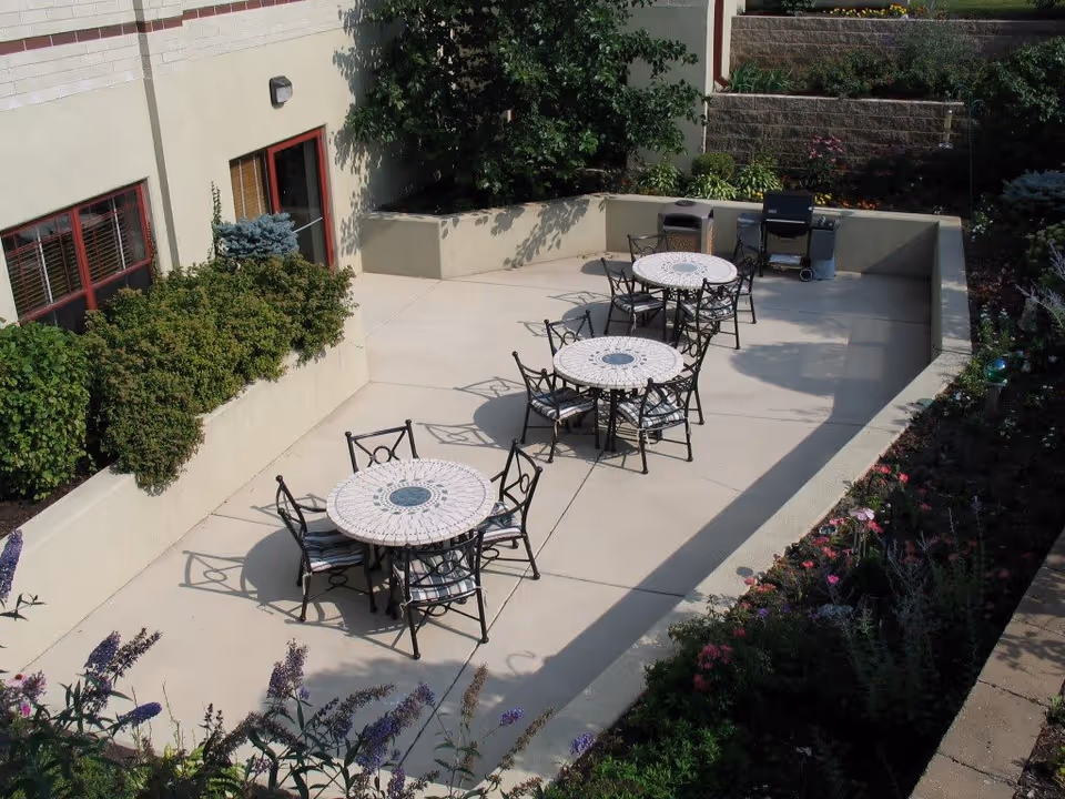 Sunlit outdoor courtyard with multiple round mosaic tables and chairs, a grill, and surrounding planted beds.