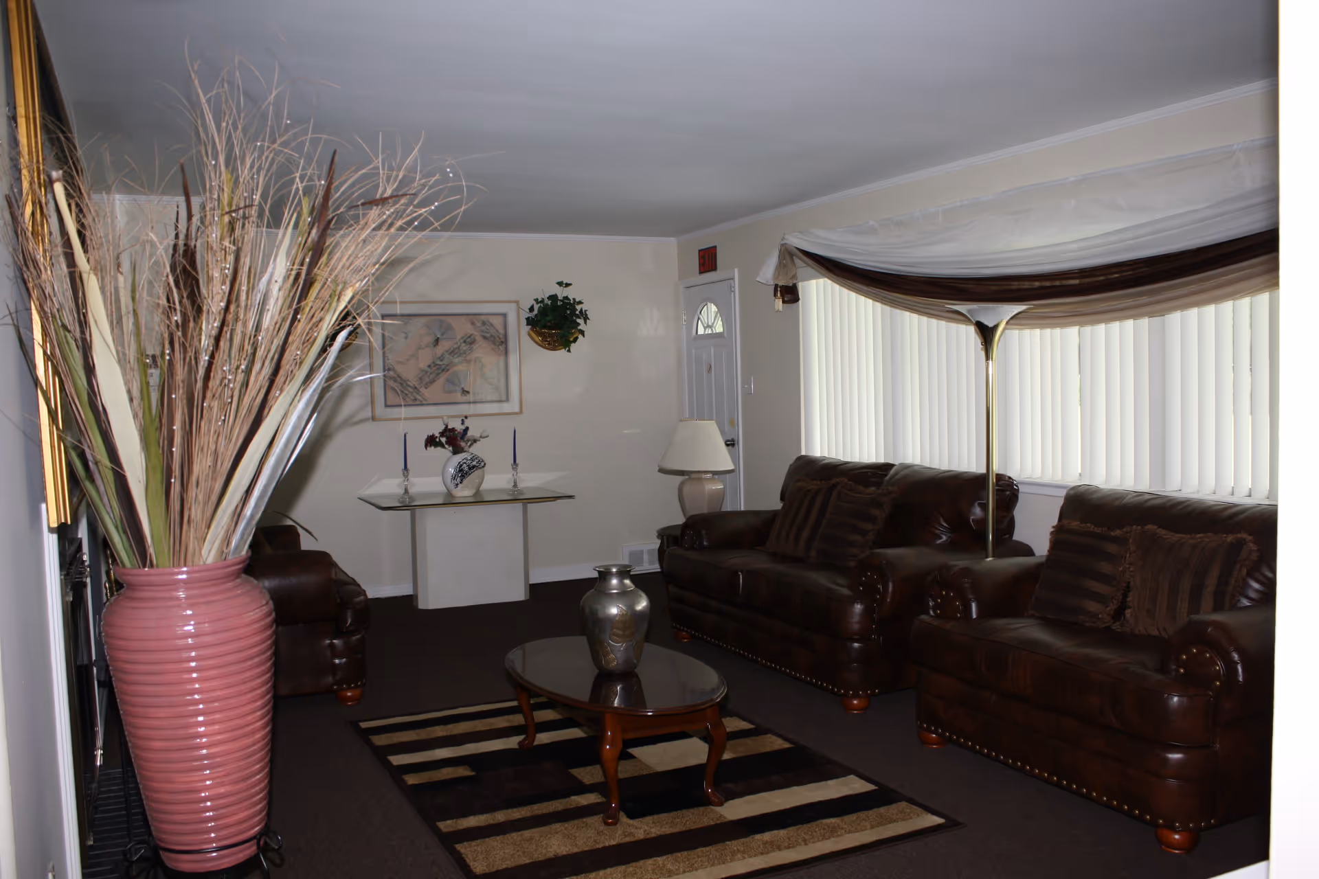 Living room with leather sofas, a coffee table, a large striped rug, and a tall pink vase of dried grasses near a window with vertical blinds.