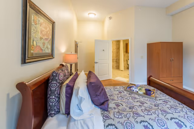 Bedroom with patterned bedding and pillows, wooden headboard, lamp, armoire, and an open door leading to a bathroom.