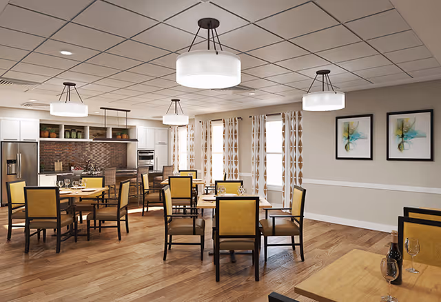 A spacious dining room with multiple wooden tables and chairs featuring yellow cushions. The room has large windows with patterned curtains allowing natural light to enter. The back wall includes a modern kitchen area with stainless steel appliances, white cabinets, and a brick backsplash. Three round ceiling lights illuminate the room, and two abstract paintings hang on the right wall.