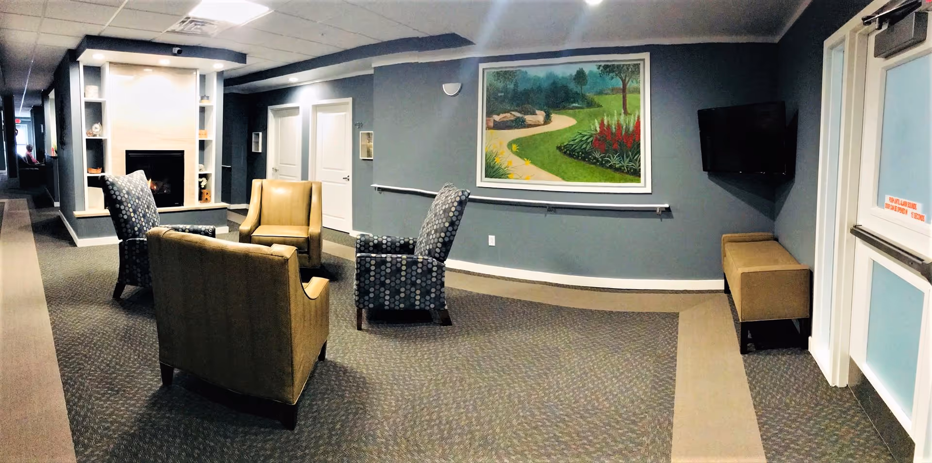 A cozy common area in a senior living facility with four armchairs arranged around a fireplace. The walls are painted gray, and there is a large painting of a garden path on one wall. A flat-screen TV is mounted on the wall near a door, and a bench is placed beneath it. The area is carpeted and has a handrail along one wall.