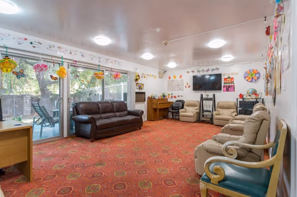 A cozy living room with a red patterned carpet, a dark brown leather sofa, several beige recliner chairs, and a wooden chair with green upholstery. The room has large sliding glass doors decorated with colorful paper flowers and butterflies, leading to an outdoor patio with chairs. The walls are adorned with children's artwork and decorations, and a flat-screen TV is mounted on the far wall above a small piano and additional chairs.