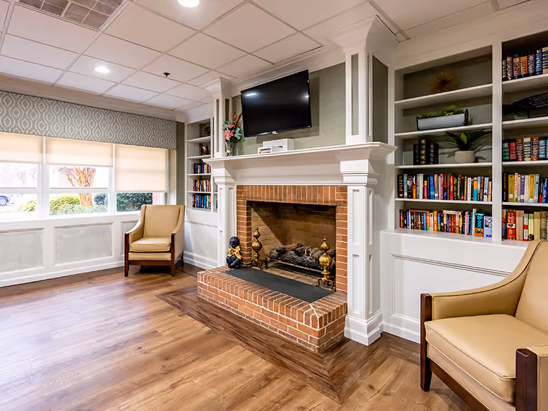 A cozy living room area with a brick fireplace in the center, flanked by built-in white bookshelves filled with books and decorative items. Above the fireplace is a mounted flat-screen TV. Two beige armchairs with wooden arms are positioned on either side of the fireplace. The room has wood flooring and a large window with beige roller shades, allowing natural light to enter.