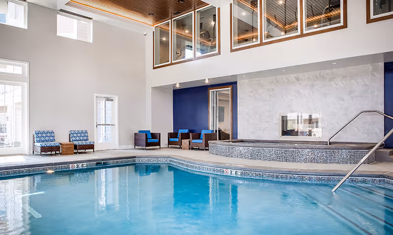 Bright indoor swimming pool and adjacent hot tub with lounge chairs and seating in a high-ceiling common area.