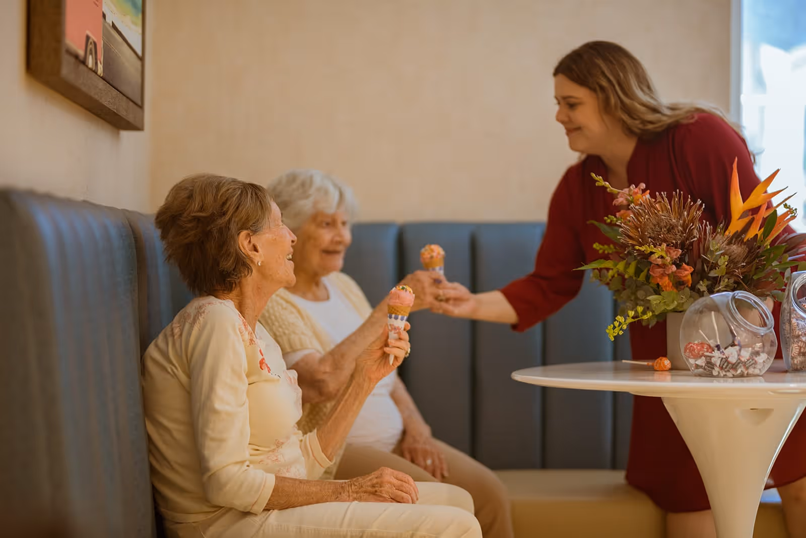 Two elderly women sitting on a cushioned bench in a cozy room, each holding an ice cream cone, while a younger woman in a red dress smiles and offers an ice cream cone to one of them. A round table nearby holds a floral arrangement and glass jars with candy.