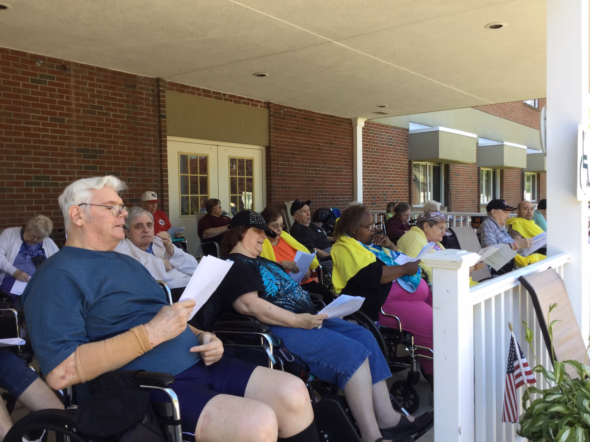 A group of elderly people sitting on a covered porch outside a brick building, many in wheelchairs, holding papers and appearing to participate in an activity together. Some are wearing yellow shawls, and there is an American flag planted in a flower pot on the porch railing.