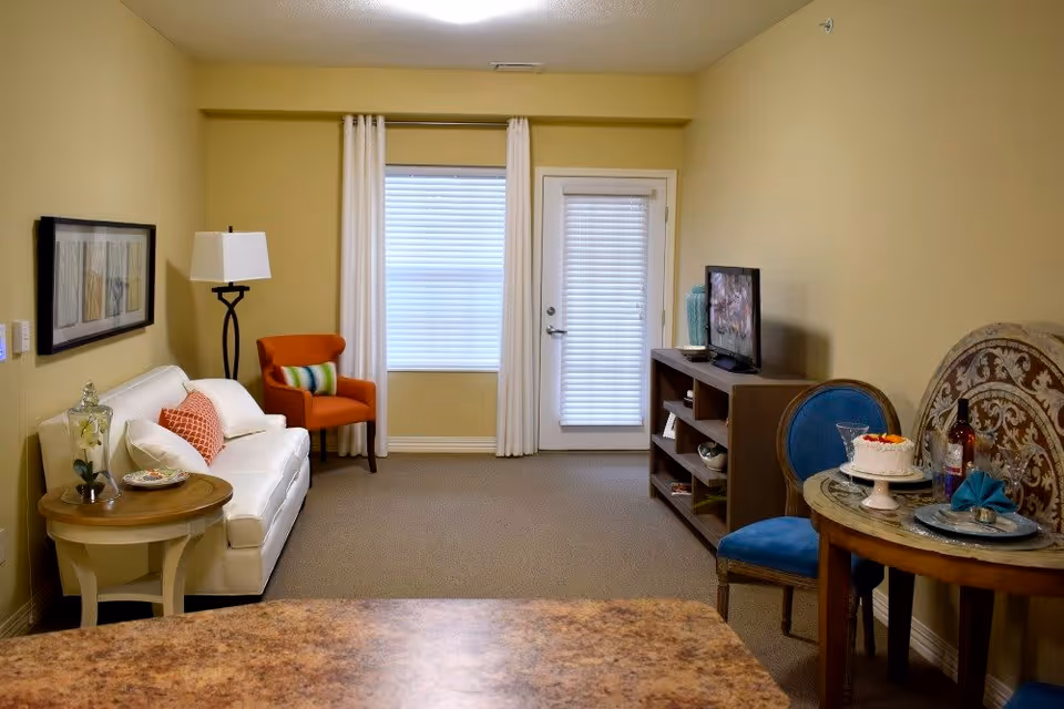 A cozy living room area with a white sofa adorned with pillows, an orange armchair with a striped cushion, a wooden side table with a glass vase and plate, a floor lamp, a window with white curtains, a door with blinds, a TV on a wooden stand, and a small round dining table set with a cake, wine bottle, glasses, and a blue chair.