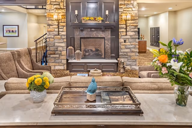 Cozy living room area with a stone fireplace in the center, flanked by two wall lamps. Beige upholstered sofas and armchairs surround the fireplace. A decorative tray with a blue bird figurine and books sits on a marble countertop in the foreground, with flower arrangements on either side. The background shows a hallway with mailboxes and a framed picture on the wall.