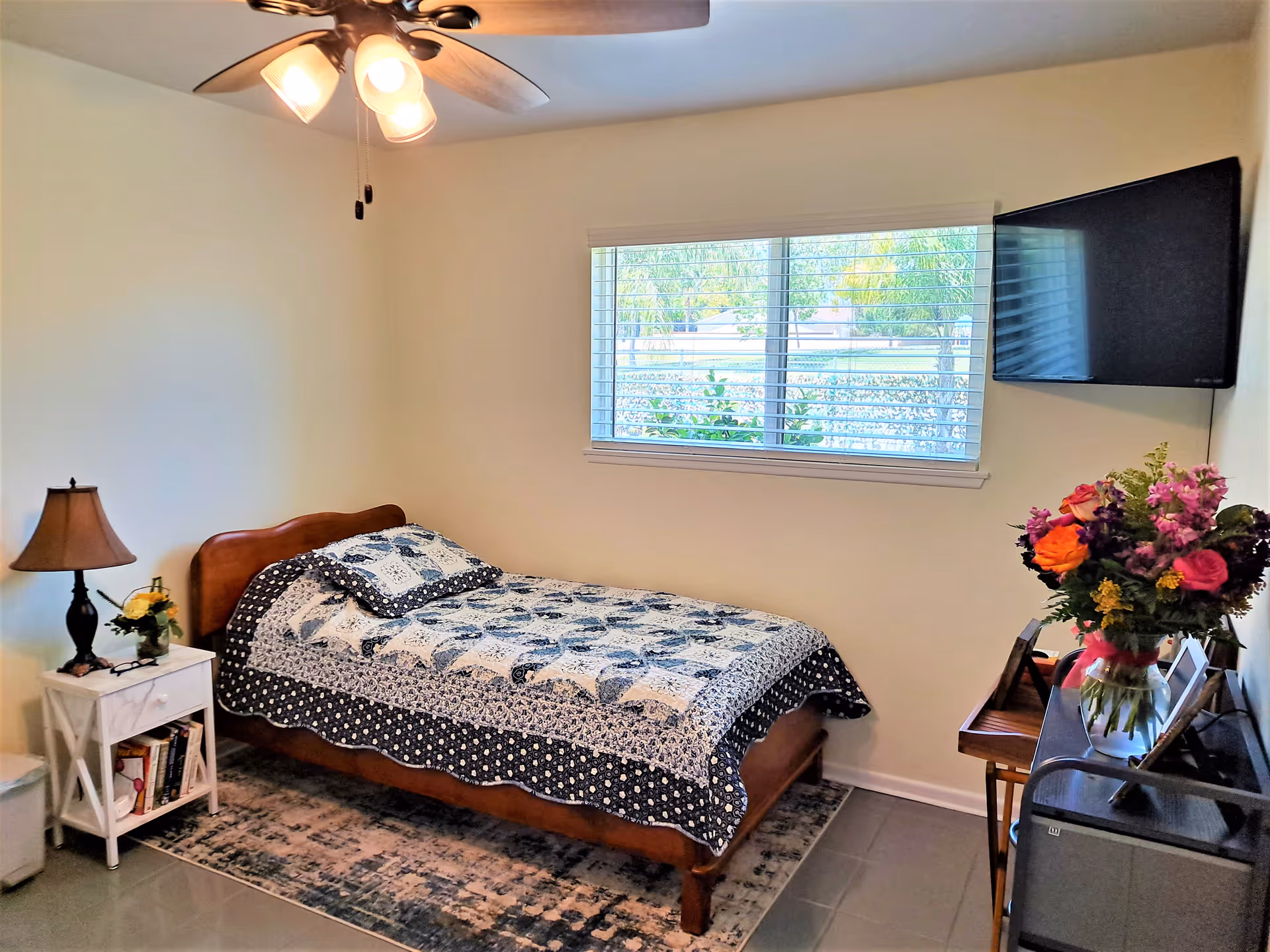 A cozy bedroom with a single wooden bed covered with a patterned quilt and pillow. Next to the bed is a white nightstand with a lamp, a small flower arrangement, and some books. A ceiling fan with lights is mounted on the ceiling. A window with blinds lets in natural light, and a flat-screen TV is mounted on the wall opposite the bed. A vase with colorful flowers sits on a piece of furniture near the TV.