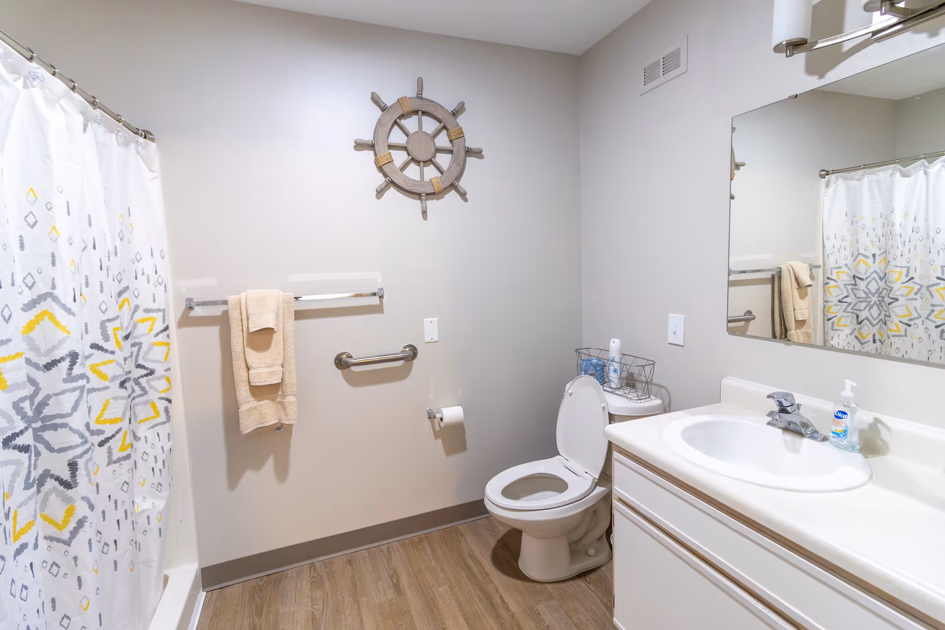 A clean bathroom with a white toilet, a sink with a countertop, a large mirror, a towel rack with beige towels, a shower curtain with a gray and yellow geometric pattern, and a decorative ship wheel mounted on the wall.
